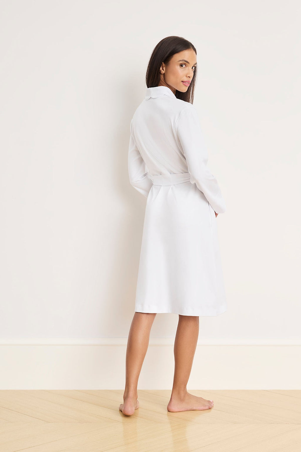A barefoot woman, wearing the Pima Scallop Robe in White made from 100% Pima cotton, with a knee-length design, long sleeves, and a scalloped lapel collar, stands on a wooden floor facing away but turns her head to look at the camera against a plain light-colored wall.
