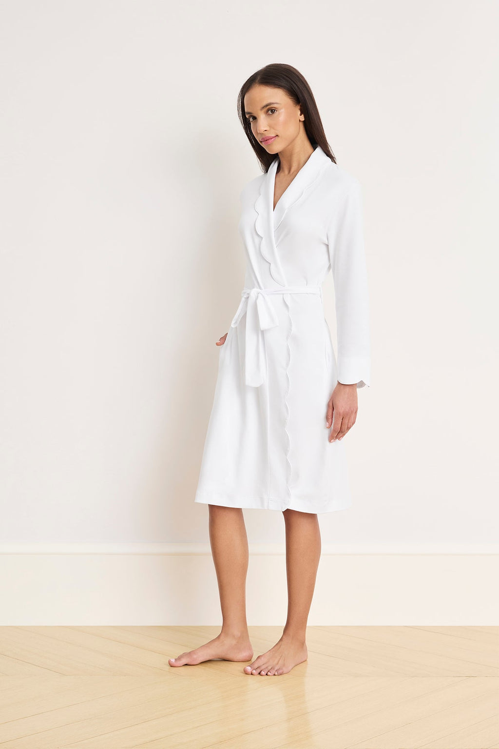 A person stands barefoot on a light wood floor, wearing the Pima Scallop Robe in White with a tie belt and scalloped lapel against a plain white wall.