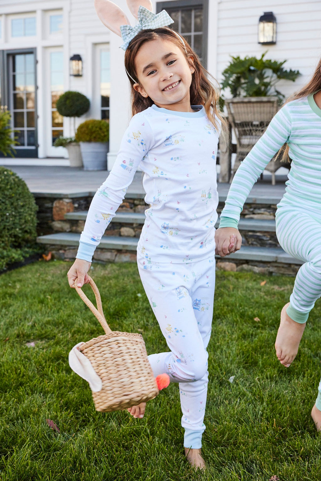 A young girl, barefoot on the grass and smiling, wears the Kids Long-Long Set in Easter Egg Hunt pajamas with a bunny ear headband and holds a bunny basket—perfect attire for an Easter Egg Hunt in front of the house.