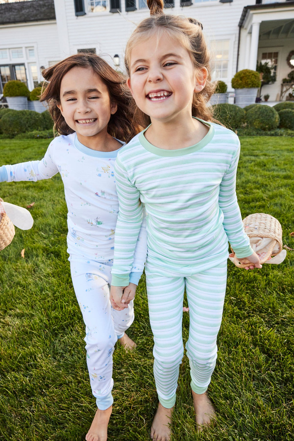 Two young girls in 100% Pima cotton pajamas smile and hold hands while walking on grass outside. One wears white pajamas with a floral print; the other wears the Kids Long-Long Set in Parisian Green Picnic Stripe. Both look happy and playful.