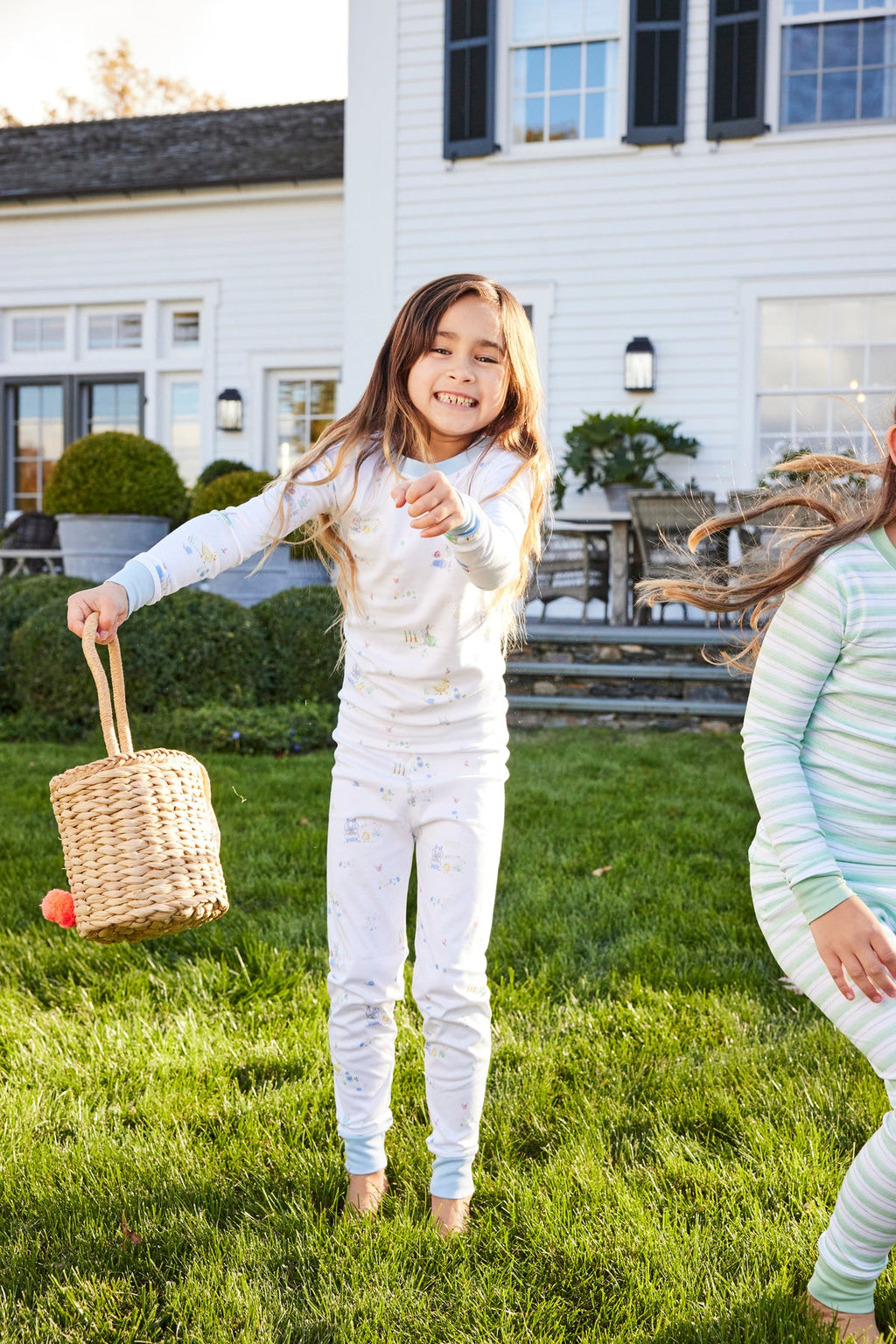 A girl in our Kids Long-Long Set in Easter Egg Hunt pajamas smiles and jumps on a grassy lawn, holding a woven basket during an Easter egg hunt outside a white house with black shutters, joined by another child.