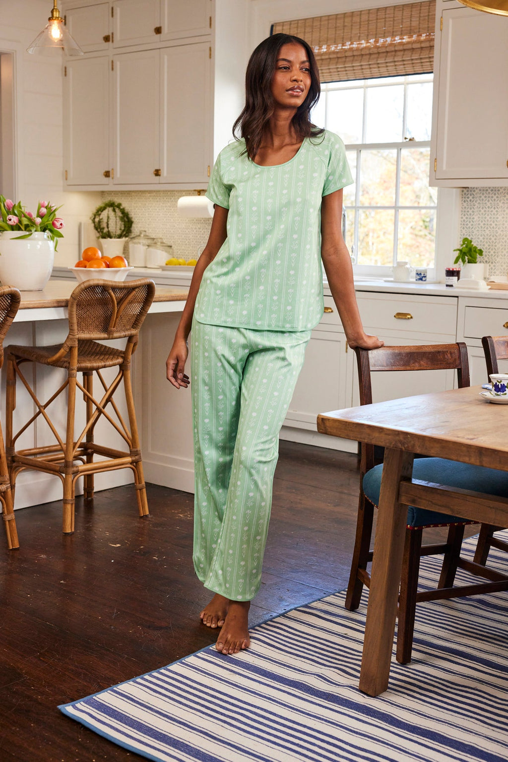 Someone wearing the Pima Short-Long Set in Parisian Green Watercolor Stems stands in a kitchen with white cabinets. The room includes a large window, wooden table on a striped rug, and barstools at the island, while fruits and plants add freshness to the counter.