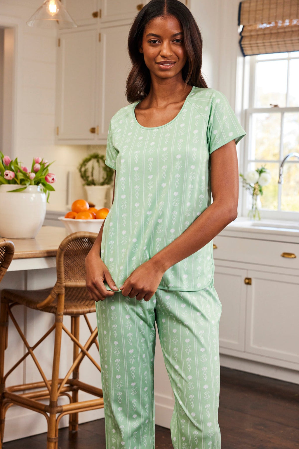 A woman in a kitchen wearing the Pima Short-Long Set in Parisian Green Watercolor Stems stands near a kitchen island with white cabinets, a window with bamboo shades, and beside her are a wicker chair, vase of tulips, and a bowl of oranges.